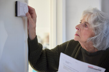 Worried Senior Woman With Bill Turning Down Central Heating Thermostat At Home In Energy Crisis