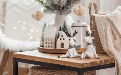 A bouquet of Christmas trees, reindeer, a blanket in a basket and Scandinavian white houses on a wooden table in the home interior of the living room. Cozy concept of festive decoration of the house.