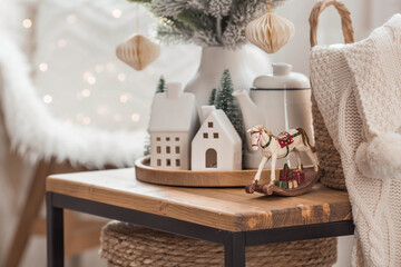 A bouquet of Christmas trees, a rocking horse, a blanket in a basket and Scandinavian white houses on a wooden table in the home interior of the living room. A cozy concept of a holiday home.