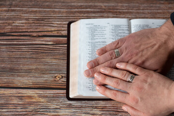 Hands of Christian couple wearing rings holding an open Holy Bible Book placed on wooden background...