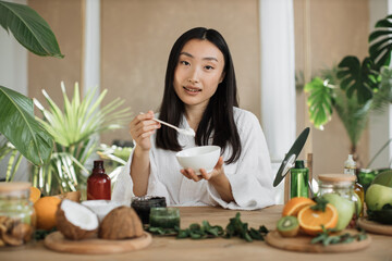 Obraz na płótnie Canvas Beautiful healthy asian woman in white bathrobe sitting at wooden table with various ingredients preparing natural cosmetics at home, mixing the mass in a bowl while preparing cream. Skin care.