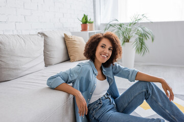 positive african american woman with curly hair sitting near modern sofa in living room.