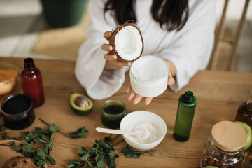 Close up hands of young woman holding half of coconut while making homemade cream for healthy skin. A lot of ingredients for home made cosmetics lying on wooden table.