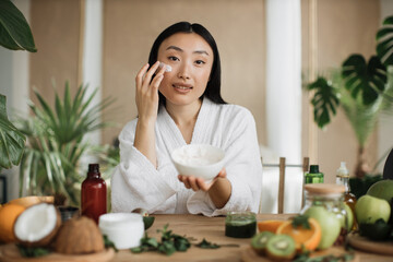 Obraz na płótnie Canvas Beautiful asian woman preparing homemade cream with natural ingredients for skin care applying on face sitting at wooden table with various organic fresh vegetables and fruits on light exotic studio