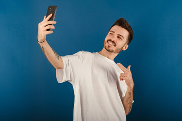 Handsome young man wearing casual clothes posing isolated over blue background pointing index finger on mobile phone while taking selfie with big smiles. Communicating concepts.