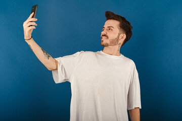 Caucasian man wearing white tee posing isolated over blue background making a selfie looking at the phone screen with big smiles.