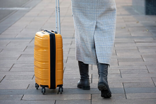 Woman in coat and boots walking with suitcase on wheels. Female legs and luggage on the street, travel concept