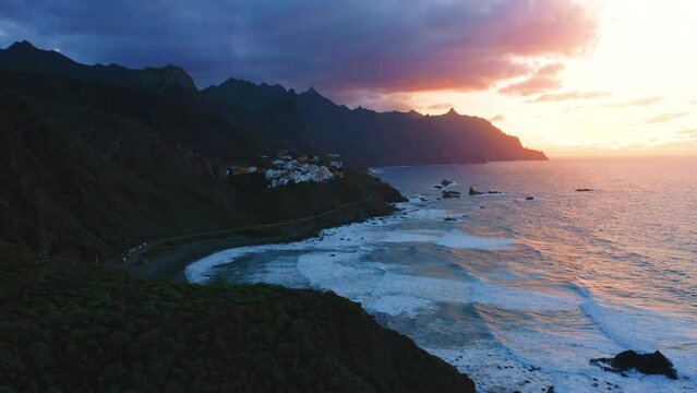Aerial view over deep dark night ocean water. Last rays of sunset on cloudy colorful sky. Little village Almaciga on top of mountain. Amazing nature landscape. Tenerife north. Canary island.