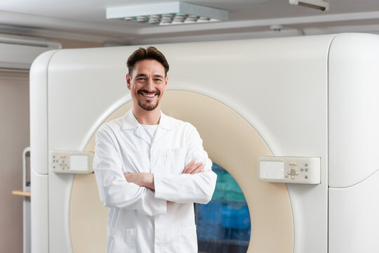 bearded physician in white coat smiling at camera near ct scanner in hospital.