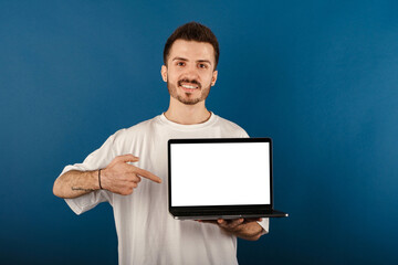 Cheerful young man wearing t-shirt posing isolated over blue background pointing with index finger to laptop pc computer with blank empty screen