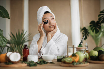 Obraz na płótnie Canvas Pretty asian woman in towel and bathrobe sitting at table with fruits, vegetables for homemade cosmetics applying patches under eyes. Charming female making beauty procedures at exotic tropical home