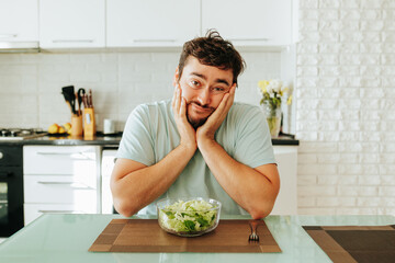 An unhappy young man with a bored look is sitting at the table and does not want to eat a salad....
