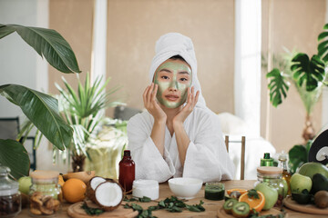 Portrait of beautiful young asian woman with green cosmetic mask on face and towel on head over...