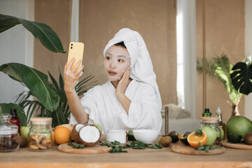Close up portrait of happy asian woman blogger holding phone applying white cotton mask sheet over exotic background. Pretty lady doing skin care procedures indoors at home, doing selfie.