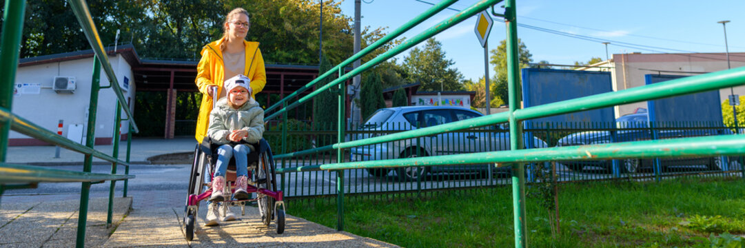 Young mother pushing a wheelchair with her daughter, girl living with cerebral palsy, on their way to therapy. Barrier free access, wheelchair or pushchair ramp.