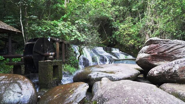 Wooden wheel of an old medieval water mill used for rice production 
 with background of a waterfall and forest.
