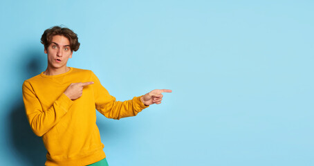 Portrait of young man with curly hair posing, showing directions with fingers isolated over blue background. One way. Flyer