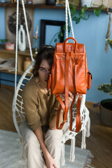 beautiful slender girl in beige pants and olive sweater posing while sitting in a swing chair with a small shiny orange leather backpack