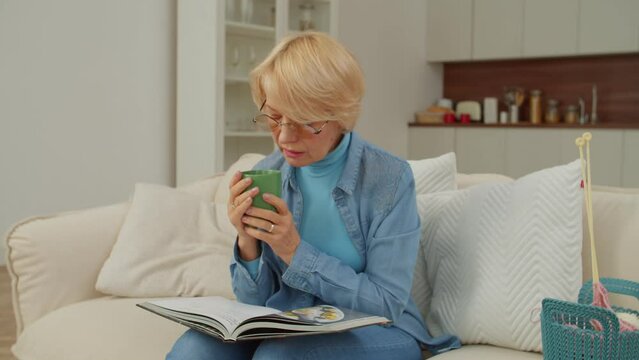 Relaxed charming retired mature female in eyeglasses sitting on sofa, sipping hot drink from cup, leafing through cookbook and looking for recipes while enjoying leisure in domestic room.