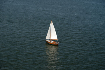 Fototapeta premium Aerial view of wooden old yacht with sail against backdrop of blue water with waves and ripples. Lonely white sail is sailing. Perfect content for posters or advertising banners, creative projects.