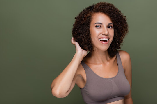 Smiling african american woman in top touching curly hair and looking away isolated on green.