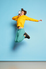 Portrait of young man with curly hair posing, shouting in megaphone in a jump isolated over blue background. News