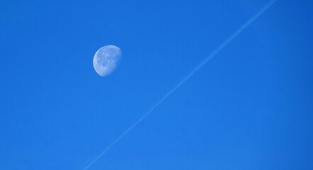 Waning gibbous moon with a jet stream in clear blue sky.