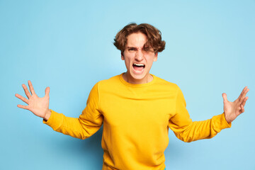 Portrait of young man with curly hair posing, emotively shouting in anger isolated over blue background