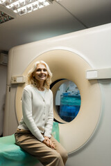 cheerful mature woman sitting near computed tomography scanner and looking at camera.