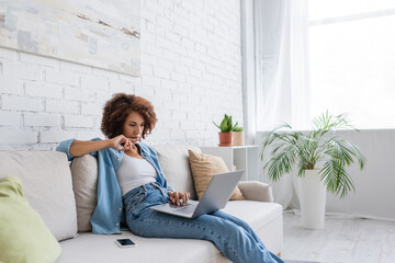 pensive african american woman using laptop while sitting on couch and working from home.