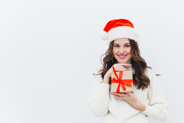 A young woman in a white sweater and a santa hat holds a Christmas present on a white background. Copy space.