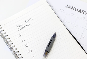 Notepad with handwritten New Year&rsquo;s resolutions list and pen on top of January calendar in the background, selective focus