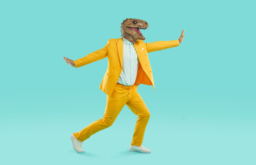 Strange man in Halloween costume dancing in studio. Side view of young guy wearing bright yellow suit and funny dinosaur mask running, dancing and swirling around on blue colour background