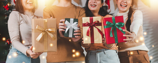 Group of young Asian man and women as friends having fun at a New Year's celebration, holding gift boxes standing by Christmas tree decoration, midnight countdown Party at home with holiday season.