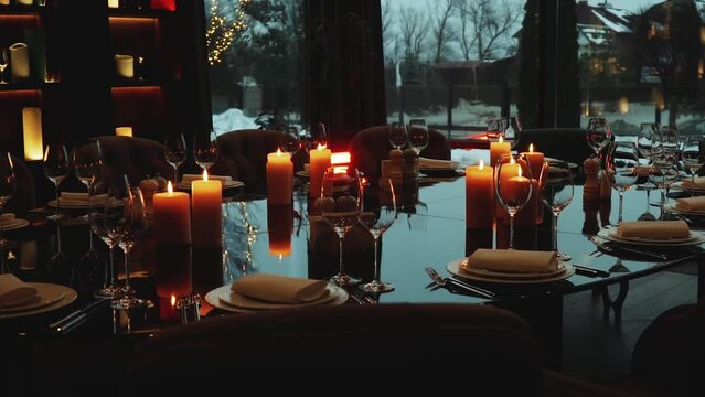 Glass glasses and white plates. Candles are burning in a dark interior of restaurant. The decor of the festive table. The plate is on a black glass table, slow motion shot.