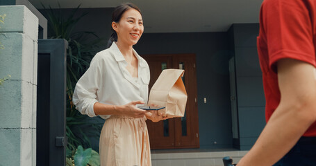 Young happy Asian woman accept paper bag from delivery man with bicycle in red uniform carry case box scan QR code payment on smartphone in front of house. express food delivery concept.