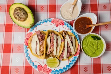 Top view of a plate of tacos next to dips and avocado, on a red checkered tablecloth