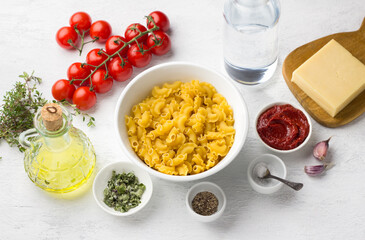 Vegetarian pasta cooking ingredients: pasta, tomatoes, cheese, olive oil, dried oregano, tomato paste, garlic, salt, black pepper, fresh thyme, water on a light gray background, top view.