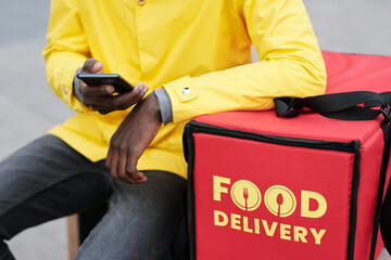 Close-up of young black man in uniform of food delivery service worker sitting by red bag with...