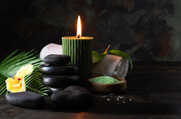 Basalt stones for massage and aroma candle on a dark table. Bath salt in a wooden bowl and a white towel in the background. Dark background with green streaks, space for text
