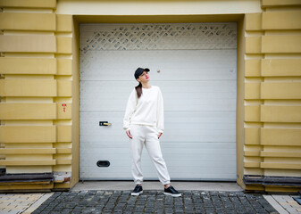 young brunette girl staying near garage doors in white sport costume, black cap and backpack