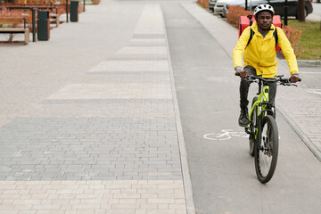 Fototapeta premium Young African American male courier with backpack riding bicycle while moving along road in park or other urban environment