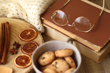 Fototapeta na wymiar Cup of tea or coffee, various spices, bowl of cookies, tangerines, books, reading glasses and knitted blanket on wooden table. Hygge at home concept, selective focus.