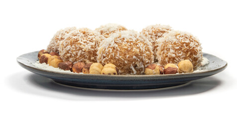homemade coconut candies with almonds inside, sprinkled with coconut chips, lying on a round plate of dark blue color, decorated with hazelnuts isolate