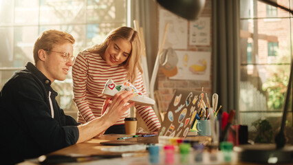 Young and Positive Female Artist Looks at Screen and Shares Ideas With her Male Colleague Who Uses Laptop Computer. Designers Creating New Model of Shoes. Modern Professionals Concept.