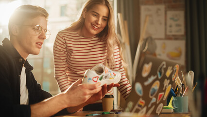 Blonde Male Designer Shares Ideas with his Female Colleague while Modelling Custom Made Sneakers in front of Laptop Computer. Professionals Create Unique Shoes. Fashion Project Concept.