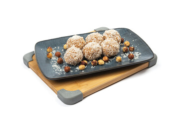 homemade coconut candies with almonds inside, sprinkled with coconut shavings, lying on a rectangular plate of dark blue color decorated with hazelnuts, standing on a bamboo board