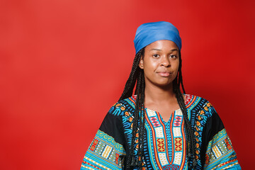 Black woman in cultural clothing looking directly into the camera. She is serious standing in front of a bright red background.