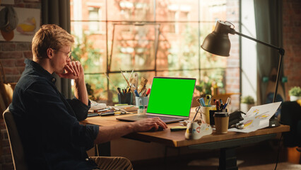 Millennial Designer Office: Caucasian Male Artist Sitting at His Desk Working on Laptop Computer With Green Screen Chroma Key on Display. E-Commerce and Smart People with Fresh Ideas Concept.