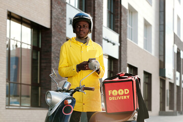 Young African American courier in helmet and uniform standing by bike with big red bag containing online food orders of customers